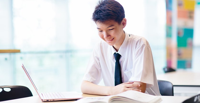 Teenager studying at desk with laptop and book